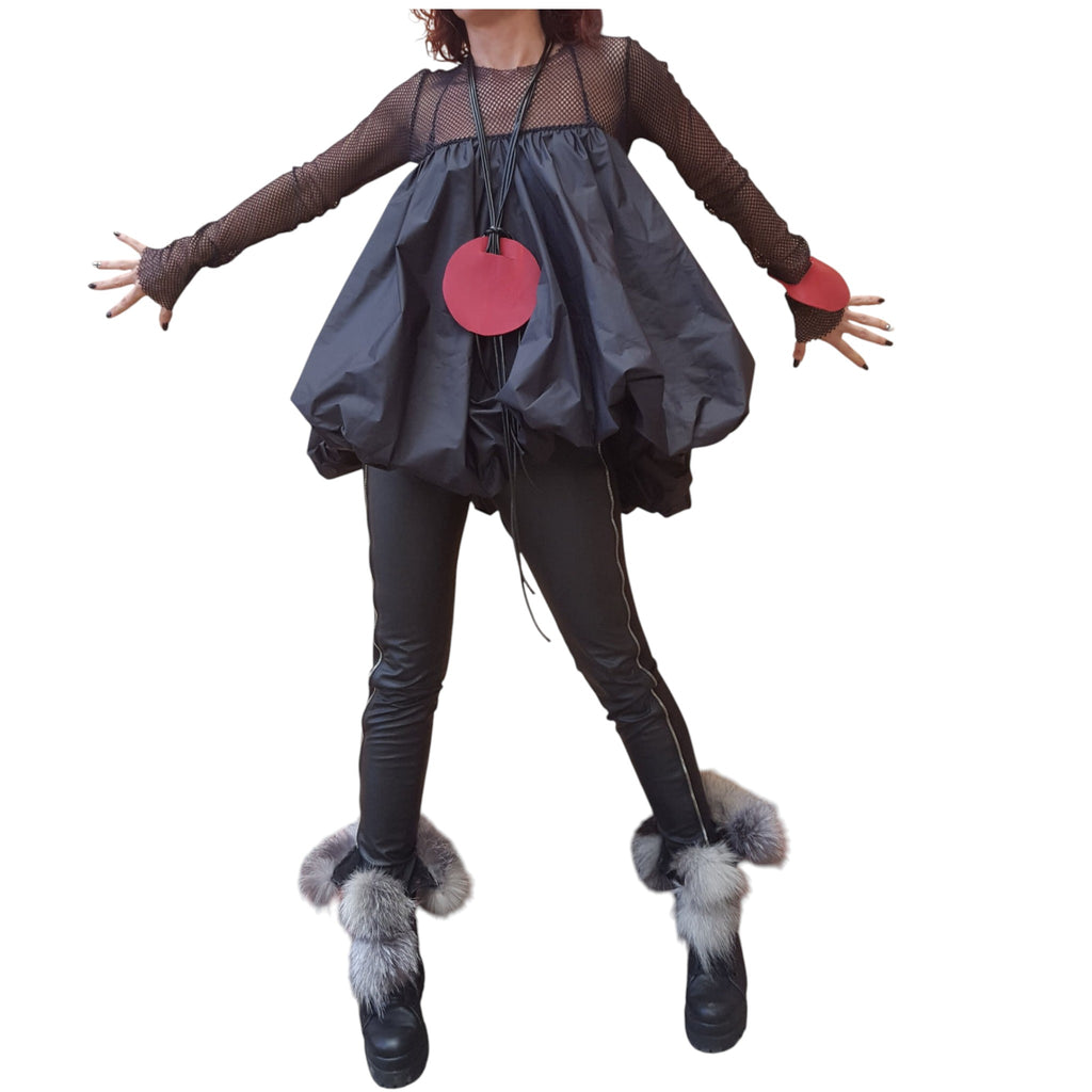 A person wearing a black outfit with a round red pendant, black leggings, and fur boots, standing with their arms outstretched.