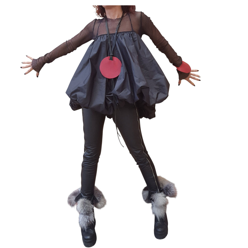 A person wearing a black outfit with a red circular pendant, black leggings, and fur boots, standing with their arms outstretched.